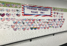 American flag hearts surround a large rectangular sign that says, "Military Veterans, Thank you" The hearts are colored in red, white and blue and are on a cinder block white wall with a green painted band at the top. There is a hand railing below the dis