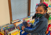 A girl from Mrs. Mkrtschjan's class is picking out a book from the colorful bins on the counter. The bins are yellow and blue. The girl is wearing a blue jacket and wearing a Panther green and black school mask. She has pink flowers in her hair. There are