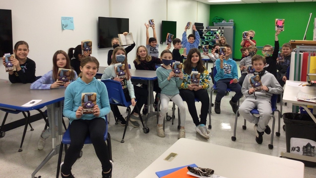 Students from Mrs. Stasi&#39;s class are sitting in chairs inside the Sullivan innovation lab located inside the library area. There are tvs on the left wall and a green painted back wall. The students are holding up books they are reading. 
