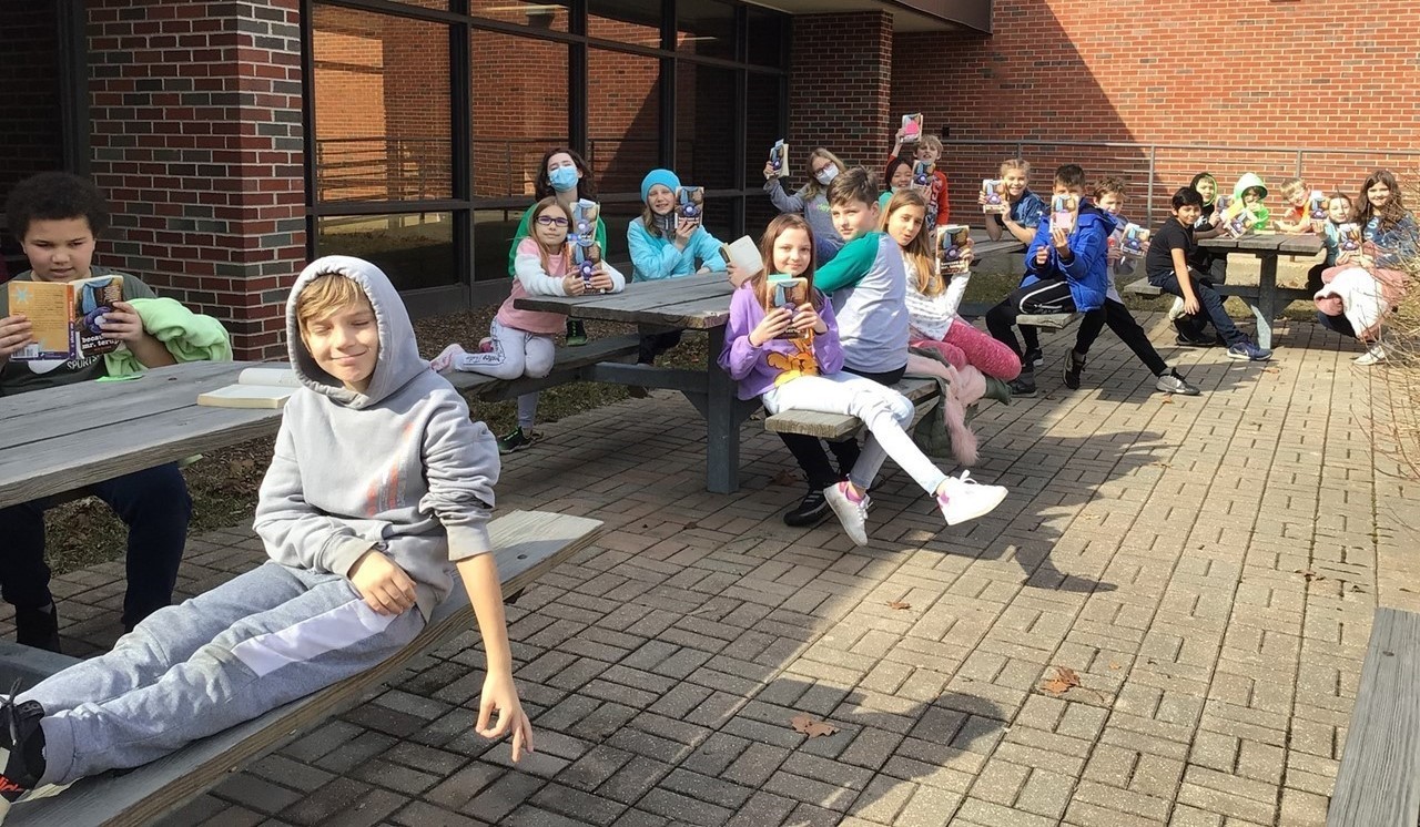 Mrs. Stasi&#39;s class is sitting outside in the sunshine outside the glass hallway connecting Ross and Sullivan at the wooden picnic tables. Students are sitting at the picnic tables holding up their reading books. In the background is a ramp to a building entrance. The school has brick walls and black borders or upper edges. There are glass window to the left and behind students. There are light tan brick pavers on the ground under the picnic tables. 