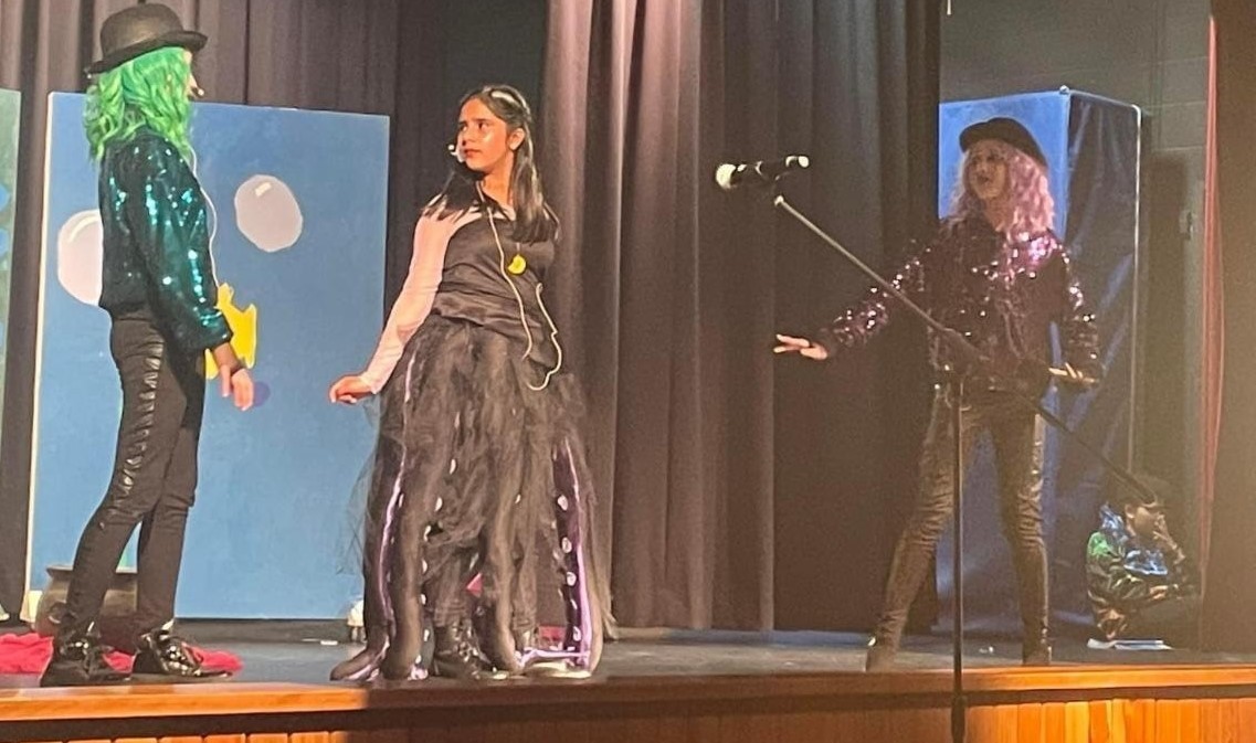 From the production of &#34;The Little Mermaid Jr.&#34; this shows Ursula wearing a black dress with long tule shaped to look like tentacles, a yellow shell necklace, and a shiny silver shirt under the dress. She has her hair down and she is looking at one of her sea serpent helpers or other characters or cast members. One eel servant of Ursula is a person on the left of her with a green wig, black top hat, shiny green shirt and black pants. The other eel character is on the right with her hands out near her hips  wearing a purple wig, black top hat, shiny purple shirt, and black pants. The eels are both looking at Ursula as they sing. There are black stage curtains behind them and in the left background you can see a stage flat showing bubbles, a yellow fish and blue background.  