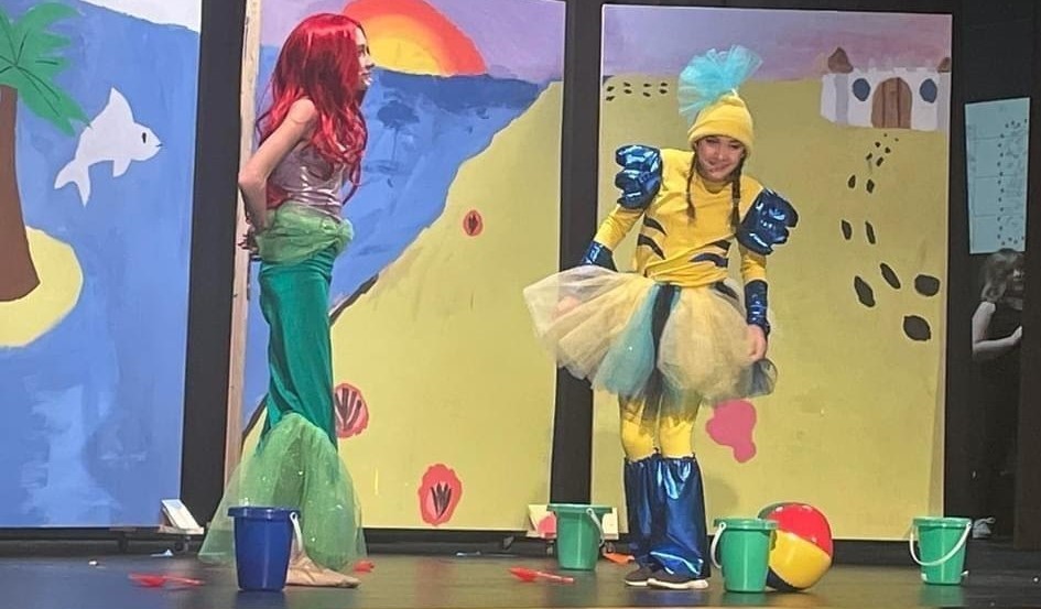 The production of &#34;The Little Mermaid Jr.&#34; shows the cast member dressed up as Ariel from the Little Mermaid. She is wearing a green skirt with tule at the bottom to look like a fin. She has a light purple top on and a red colored long wig. Her fish friend Flounder is wearing yellow and blue with tule skirt and a yellow hat with blue tule that looks like a fin on her head. The background has three flats that looks like a sandy yellow beach with a building in the background. The ocean is blue with a setting sun over the ocean. In the ocean has a dolphin jumping out of the water and an island with one palm tree on it. 