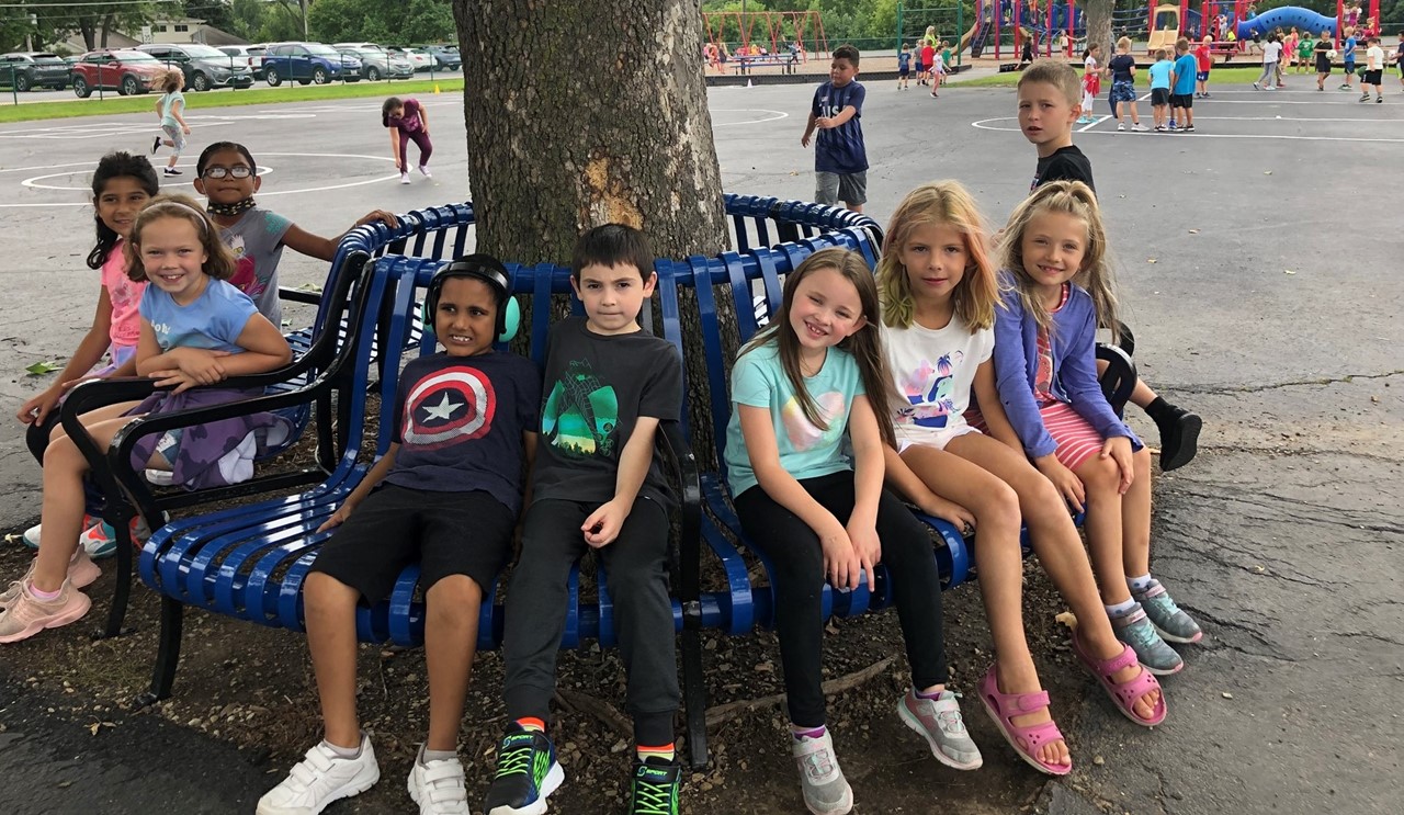 Students sitting on a round bench under a tree at recess.